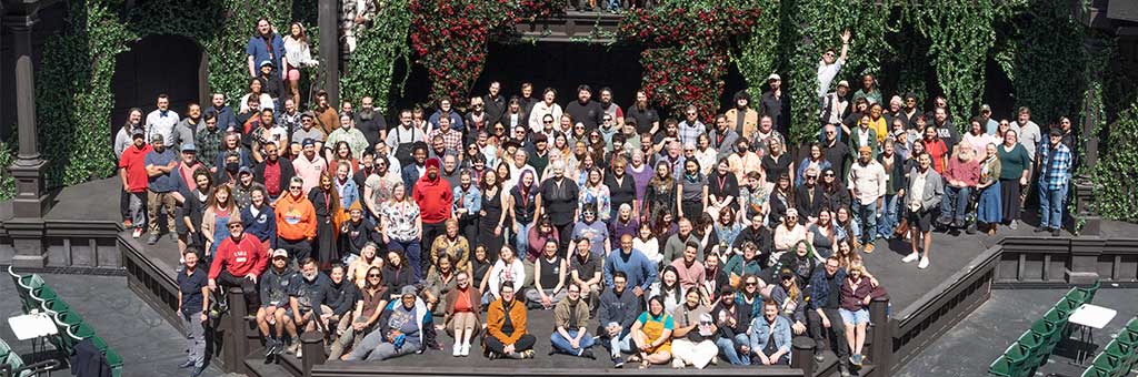 OSF company members gather on an outdoor stage with ivy-covered walls, arranged in multiple rows, posing for a group photo.