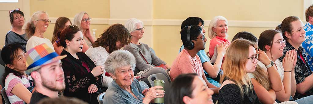 A diverse audience sitting attentively in a room, some smiling and clapping. The group includes people of various ages, and one individual is wearing headphones, while another has a colorful hat.