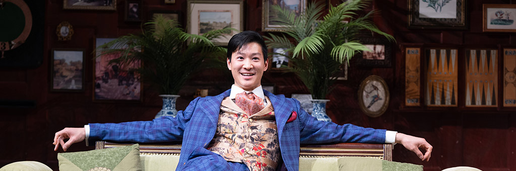 A performer in an ornate blue suit lounges confidently on a green sofa on a stage set decorated with framed art and plants, smiling at the audience.