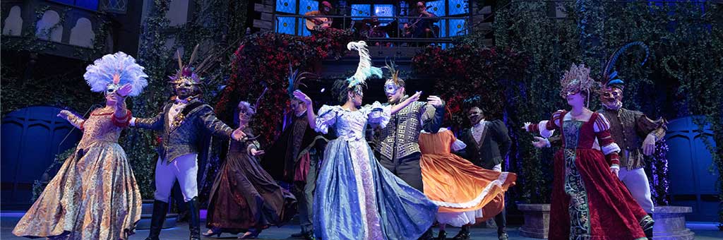A group of performers in elaborate Renaissance-style costumes and masks are dancing in a grand, colorful ballroom scene. The backdrop is decorated with ivy and a balcony where more performers are present. The image conveys a festive and theatrical atmosphere.