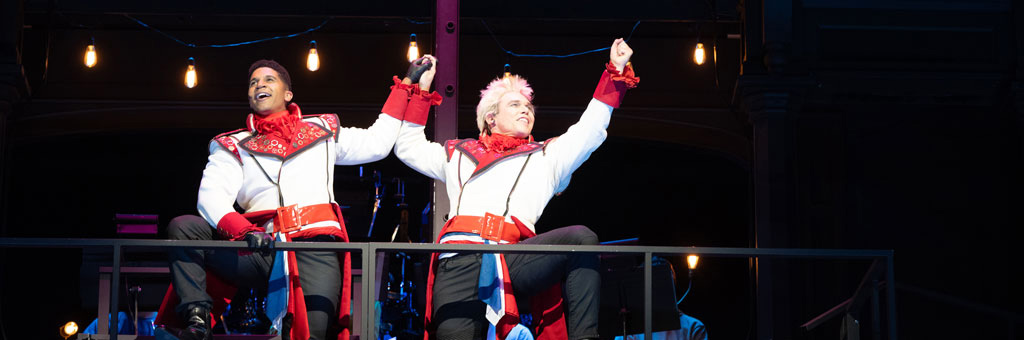 Two performers in matching costumes raising fists in triumph on stage balcony.