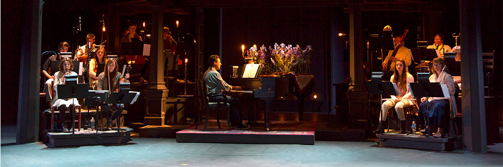 A scene from OSF's 2014 production of Into the Woods, featuring the orchestra on stage with musicians playing various instruments. The centerpiece is a pianist seated at a grand piano decorated with vibrant purple flowers, surrounded by warm, atmospheric lighting.