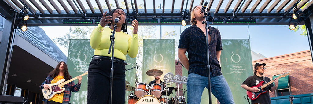 Two people in the front of a stage singing into microphones with three behind them playing intruments.