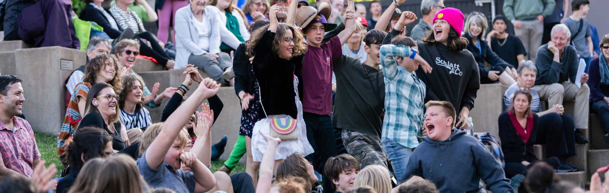 Large group of people sitting on outdoor steps cheering with raised arms during a lively community event. Several children stand in the foreground celebrating while others smile and clap in the background.