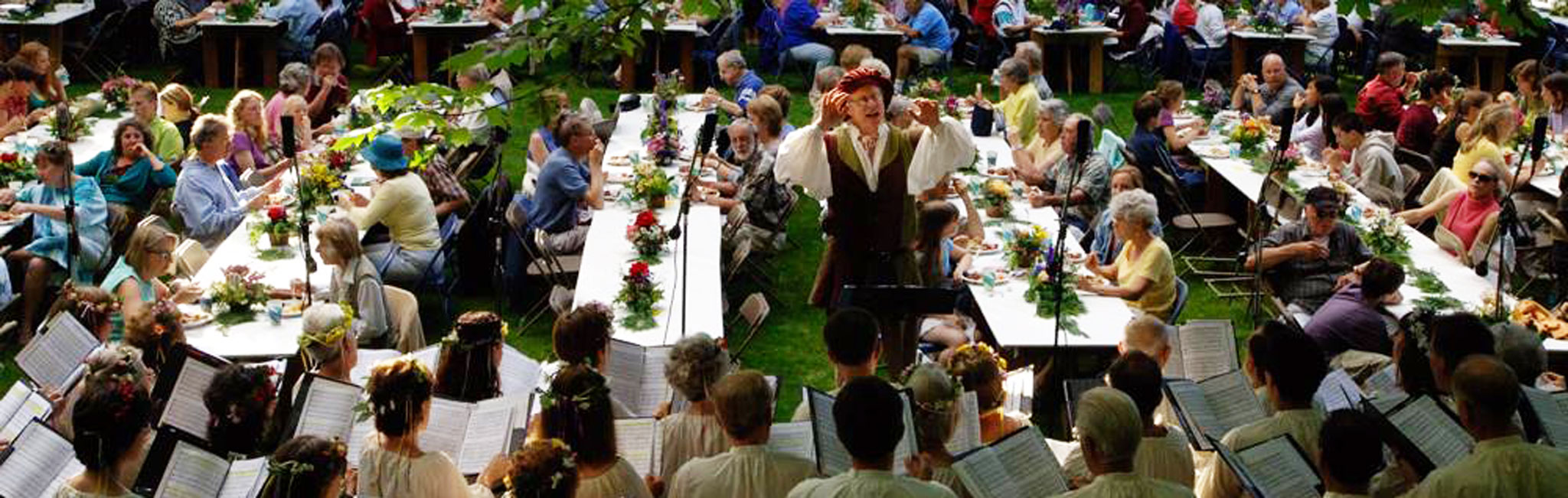 Many tables set with white table clothes and flower arrangments. Dozens of people sit around the tables.