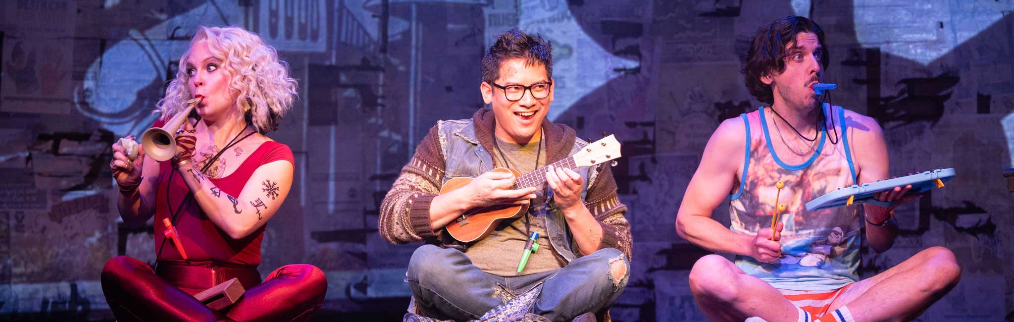 Three performers sit cross legged on a stage playing small toy instruments including a trumpet a ukulele and a xylophone.