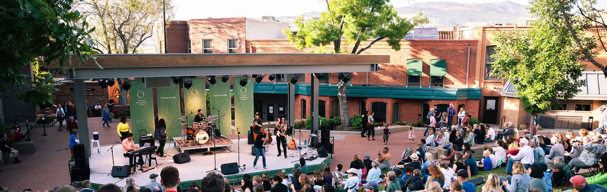 A lively outdoor stage performance at the Oregon Shakespeare Festival’s Green Show, with musicians playing instruments and an audience seated on grass and steps in a sunlit courtyard surrounded by brick buildings.