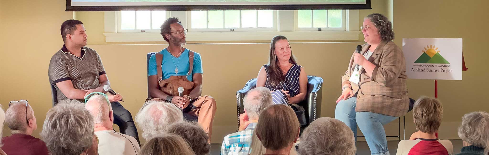 Four panelists sit on stage in front of an audience, each holding microphones. A sign behind them displays "Ashland Sunrise Project," indicating the event theme.