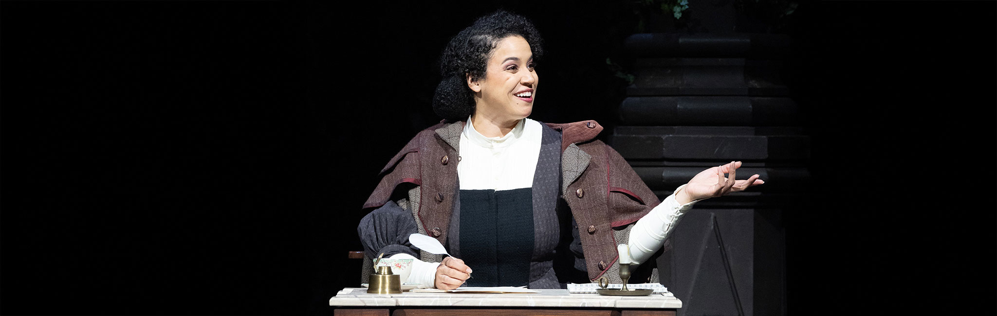 Person in a brown and gray period costume sits at a wooden desk with an inkwell and papers, smiling and gesturing to the right. Dark background with a stone pillar suggests an indoor setting.