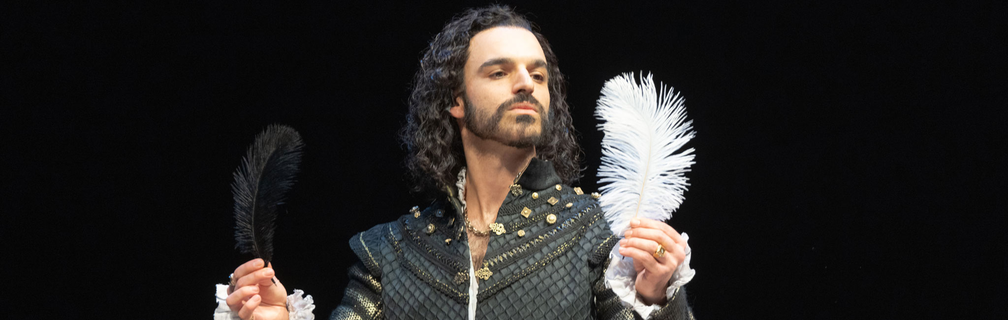 A performer in an ornate black costume with gold details holds a black feather in one hand and a white feather in the other, striking a dramatic pose against a dark background.