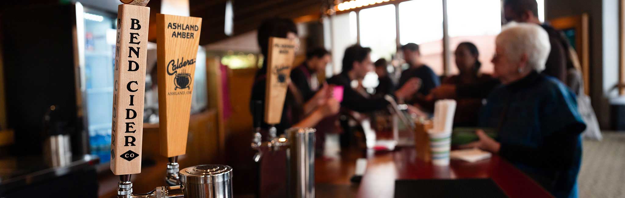 Close-up of a cider tap labeled "Bend Cider Co" alongside another tap labeled "Ashland Amber Caldera Brewing Company," with people blurred in the background a concession stand. The scene conveys a social atmosphere with patrons interacting while ordering drinks.