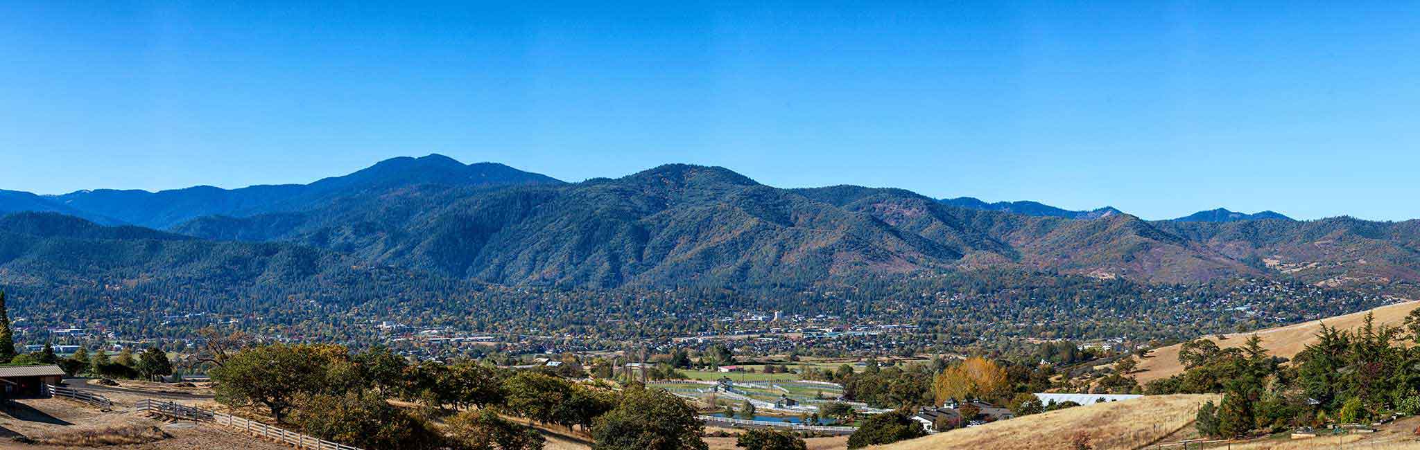 A panoramic view of Ashland, Oregon, with a valley town nestled among forested mountains and rolling golden hills. The bright blue sky contrasts with the lush greenery and earthy tones of the landscape.