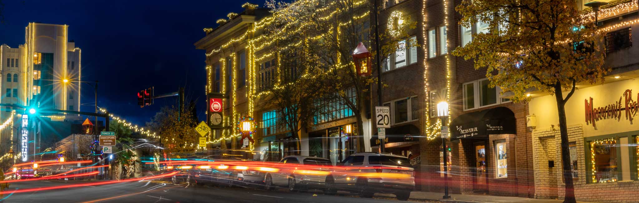 Downtown street at night adorned with festive string lights on buildings, showcasing a clock tower and vibrant storefronts. Blurred light trails from passing cars add dynamic motion to the scene.