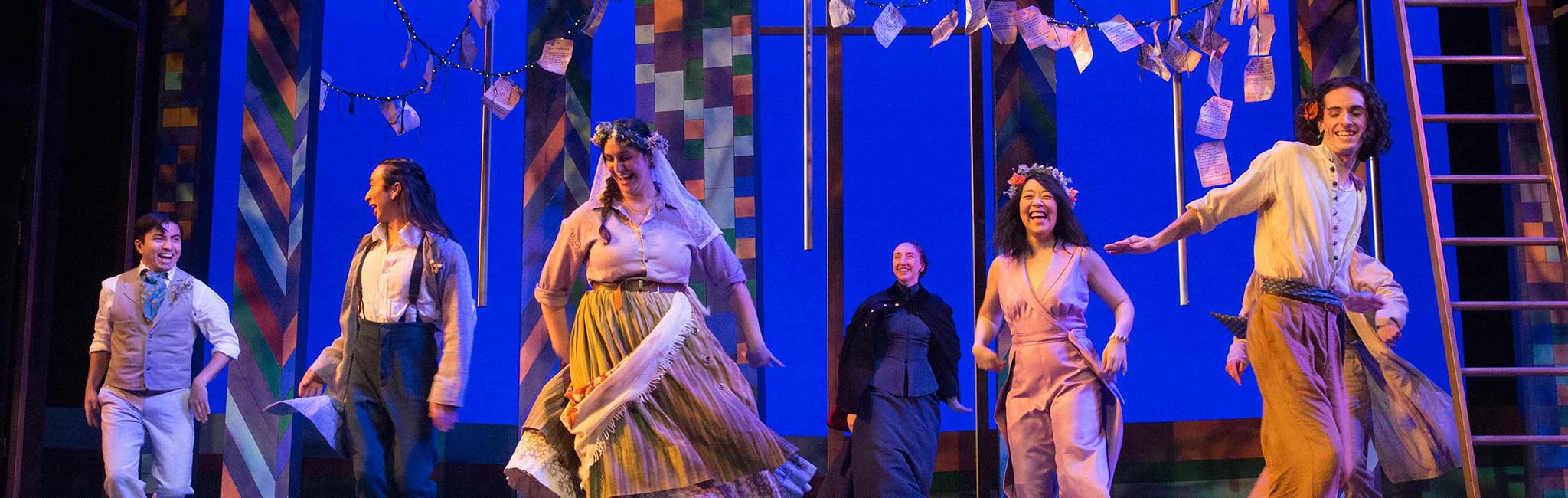 A group of performers on stage in colorful period-style costumes, joyfully dancing and interacting. The stage features a vibrant geometric backdrop, hanging string lights, and floating pages, creating a whimsical atmosphere. The actors' expressive faces and movements convey a lively, celebratory scene.