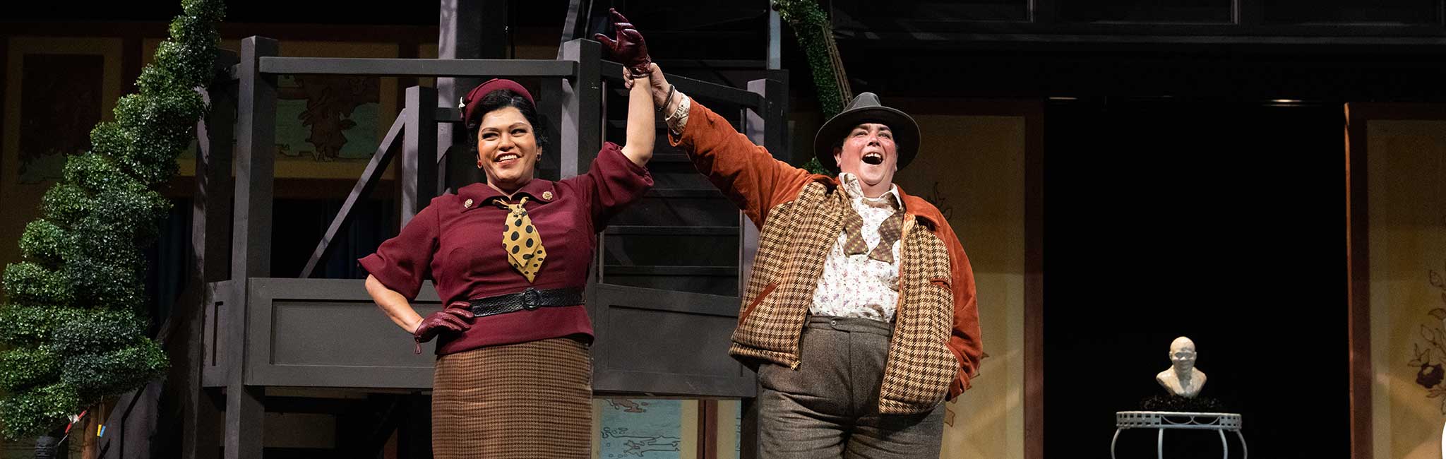 Two performers on stage hold hands in the air, smiling triumphantly. One wears a vintage burgundy outfit with a hat and gloves, while the other sports a tweed vest, loose shirt, and wide-brimmed hat.