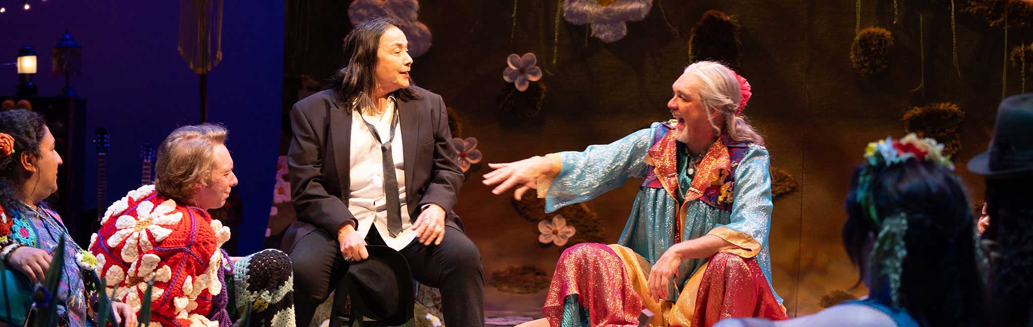 Actors sit in a circle onstage during a colorful theatrical performance. One performer in a sparkling blue and pink robe gestures animatedly while another in a black suit listens intently.