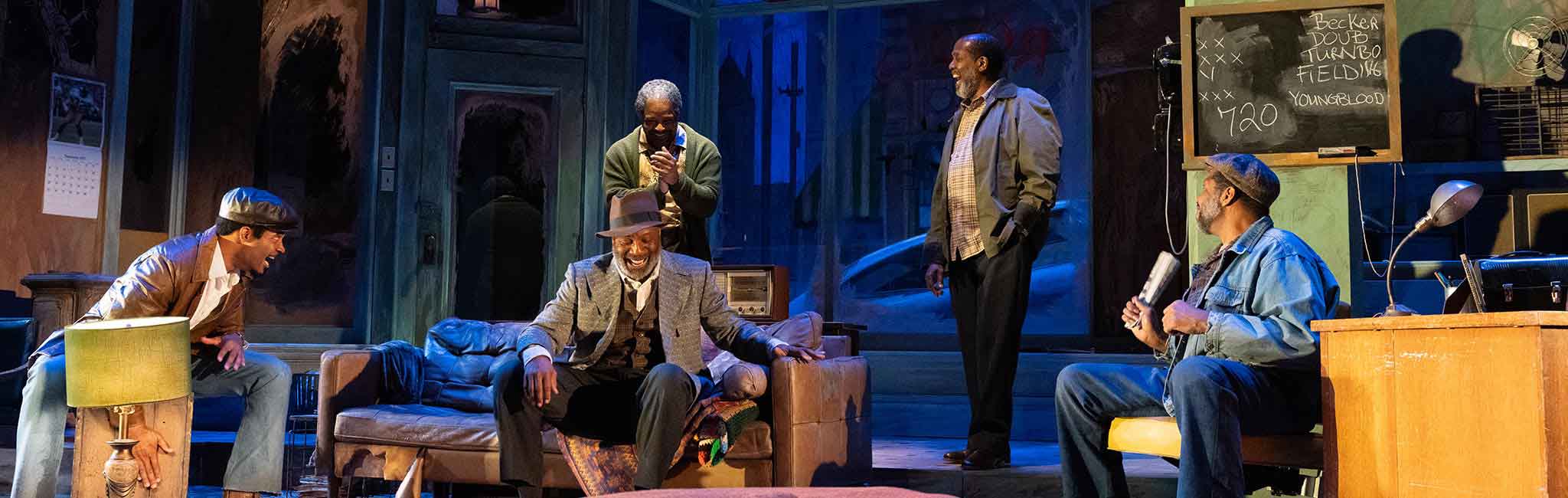 Stage scene inside a cozy living room where five actors gather around a couch, laughing and talking.