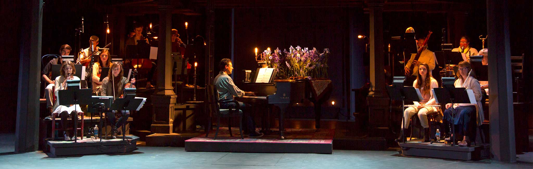 A scene from OSF's 2014 production of Into the Woods, featuring the orchestra on stage with musicians playing various instruments. The centerpiece is a pianist seated at a grand piano decorated with vibrant purple flowers, surrounded by warm, atmospheric lighting.