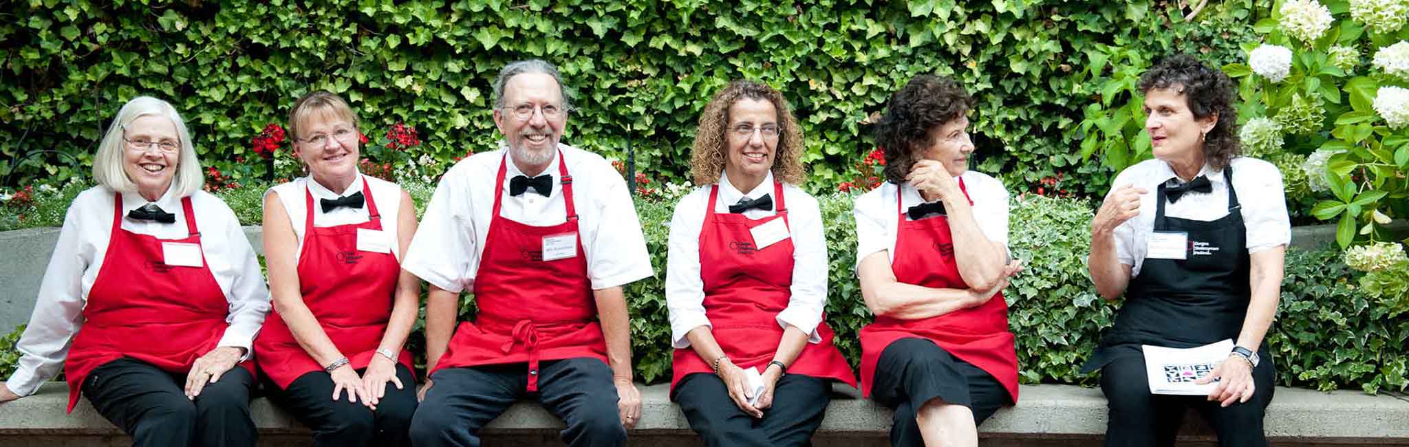 Six volunteers sitting outside on a low wall wearing aprons. 