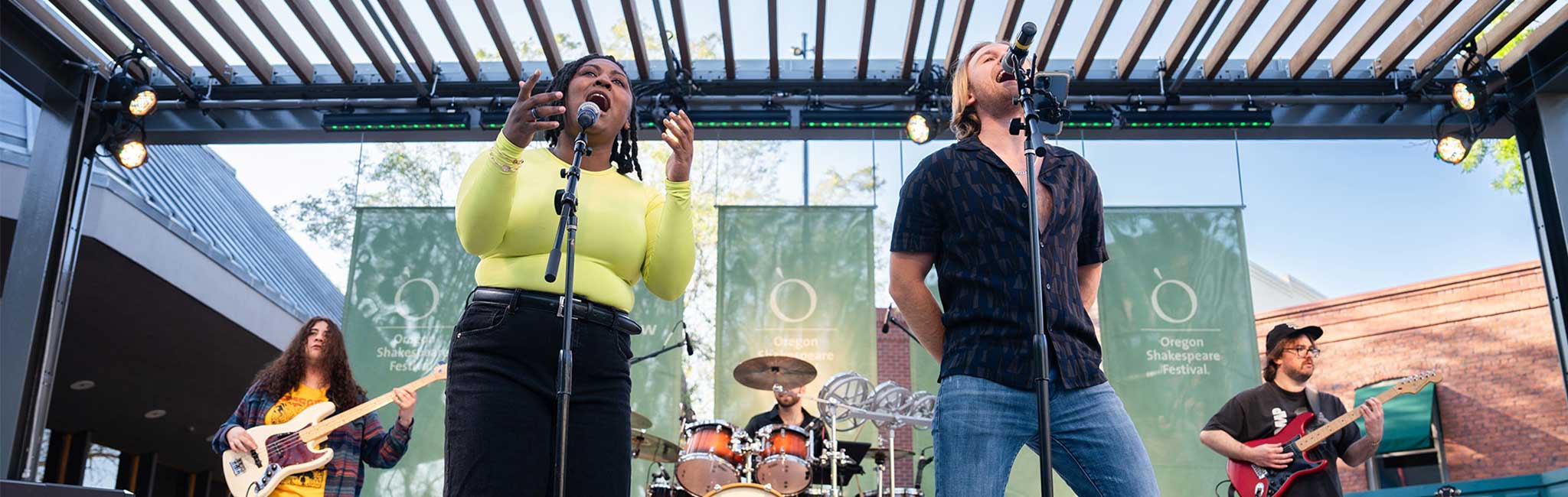 Two people in the front of a stage singing into microphones with three behind them playing intruments.